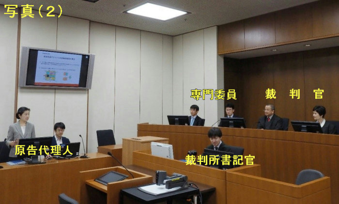 写真:(2)原告代理人がプレゼンテーションを行っている様子です。原告代理人席の奥に設置された壁掛けモニターに資料を映しながら、原告代理人が起立し説明しています。