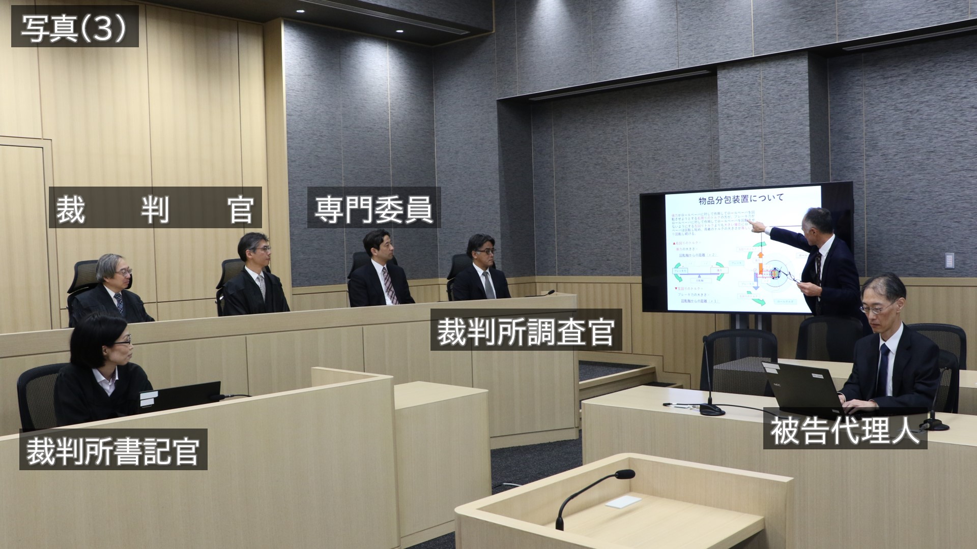 写真:写真(3)被告代理人がプレゼンテーションを行っている様子です。被告代理人席の奥に設置された壁掛けモニターに資料を映しながら被告代理人がモニターを指しながら説明をしています。
